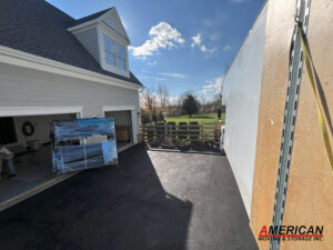House with gray siding and dormer window; open garage reveals wrapped furniture on a dolly in the driveway next to a moving truck, on a sunny day