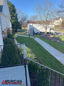 Two movers carry a wrapped item toward a white moving truck in a suburban front yard as a black metal railing and a stone path frame the scene.
