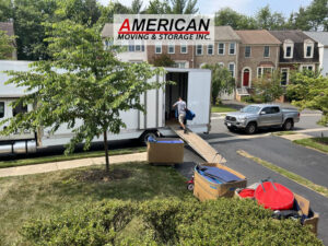 Moving company truck with ramp unloading boxes into a suburban home, logo visible on the truck.