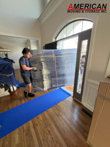 Two movers carry a large wrapped object through a doorway while a blue moving mat lies on the wooden floor; a plastic cover secures the item as they work.