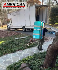 Mover from American Moving & Storage Inc carrying a large turquoise plastic bin toward a white moving truck in a suburban driveway, strapped securely to the worker's back guard.