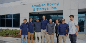 Group of eight employees wearing navy shirts and casual clothes pose together outside the American Moving & Storage, Inc. building.