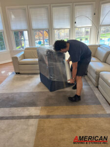 Mover in a dark blue shirt wraps a large couch with plastic stretch wrap in a bright living room.