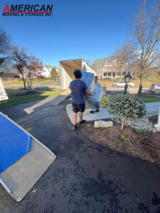 Moving crew loading a wrapped sofa into a moving truck in a suburban driveway; logo for American Moving & Storage Inc visible in the sky.