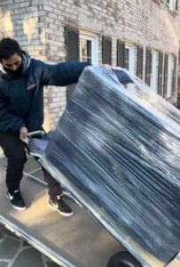 Delivery worker wearing a mask pushes a large item wrapped in plastic on a dolly along a sidewalk outside a brick apartment building