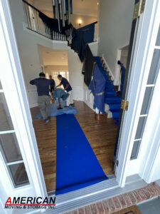 Front door opens to a two‑story foyer where movers wrap items in blue moving blankets near a staircase, with a blue rug on the hardwood floor and the American Moving & Storage logo in the corner.