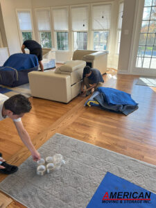 Three movers in a sunlit living room wrapping a sofa and preparing furniture for a move, with blankets and pads on the floor.