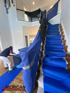 Mover wrapping a blue protective blanket around a wooden staircase in a two‑story foyer for a move; American Moving & Storage logo visible.